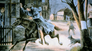 A man in a blue coat rides a dark horse through a town.