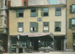 Paul Revere House with yellow siding and green window frames and shutters. Retail space on the ground floor with a threadbare red and white striped awning.