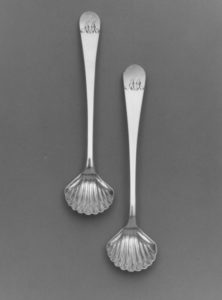 Two silver salt spoons. A seashell design on the bowl of the spoon and a letter engraving on the handle adorn the items.