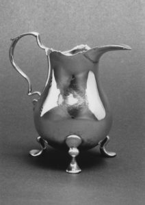 Silver cream pot with three legs, long spout, and curved handle.