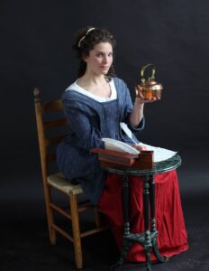 Judith Kalaora in costume as Rachel Revere, holding a copper tea kettle