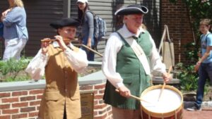 a person playing a drum next to a person playing a fife, both in colonial dress