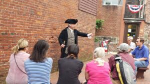 man in fancy colonial clothing gesturing to a crowd of people on benches