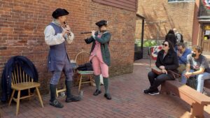 Two musicians dressed in 18th century clothing playing wind instruments in front of a brick wall. Some onlookers sit on the right side of the image.