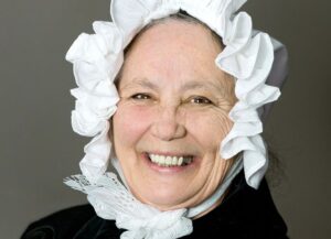 A middle-aged white woman in a white mobcap and black clothing, smiling at the camera, shown from the shoulders up.