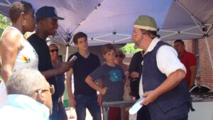 A man on the right of the image faces a group of people, chatting and demonstrating something with metal.