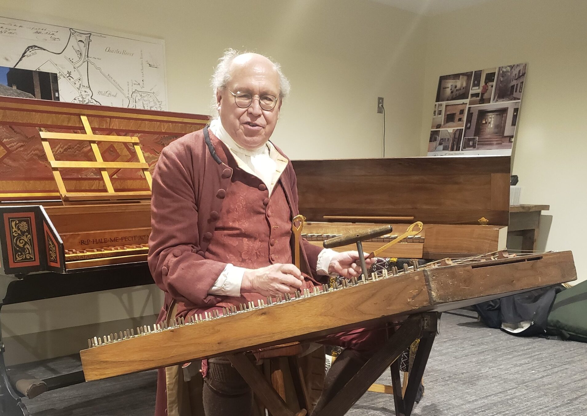 A middle-aged white man in a red colonial-era jacket and waistcoat playing the hammered dulcimer