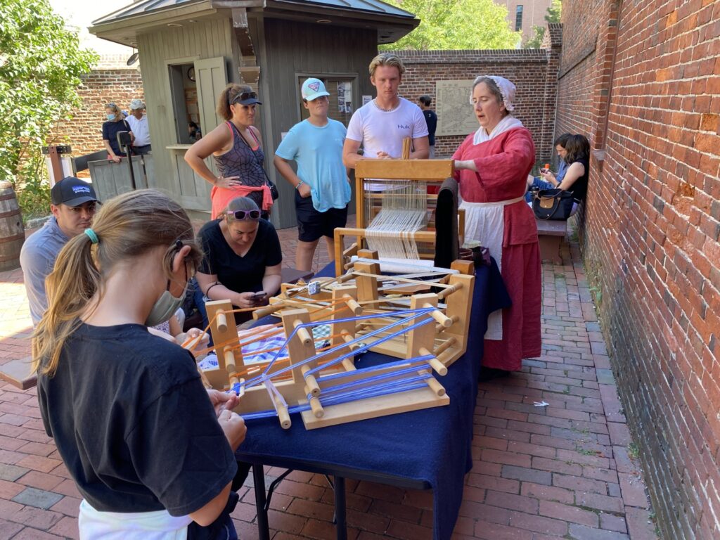 visitors at the Revere House obseving a weaving demonstration by a woman in colonial dress